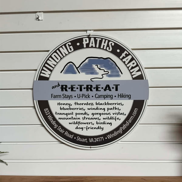 Circular metal sign for “Winding Paths Farm” and “R-E-T-R-E-A-T” mounted on a wooden wall, featuring rustic design elements and clear, bold lettering.