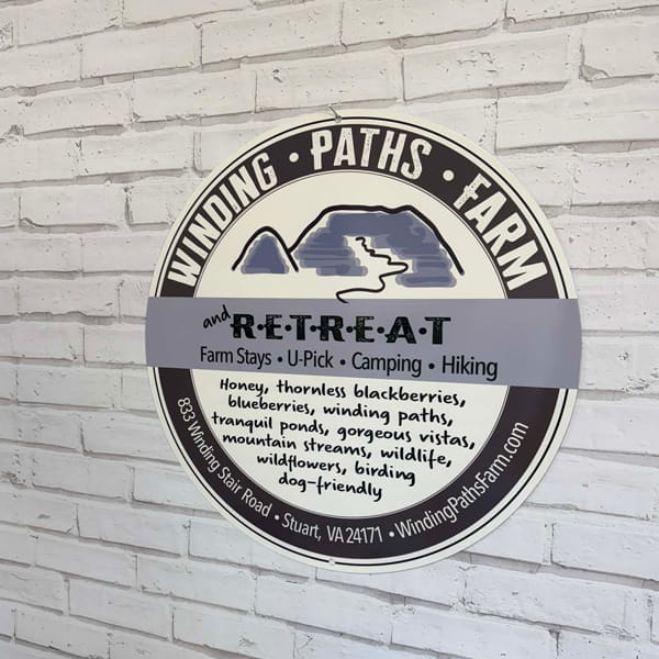Circular metal sign for “Winding Paths Farm and Retreat” mounted on a white brick wall, featuring rustic design elements and bold, easy-to-read lettering.