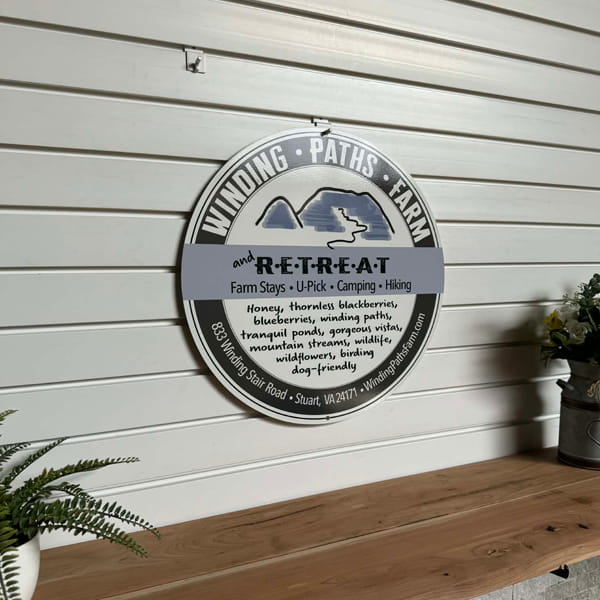 Round metal sign for “Winding Paths Farm and Retreat” displayed on a wooden surface with surrounding plants, highlighting rustic design and decorative outdoor appeal.