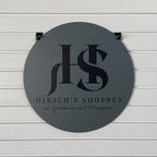 Round metal sign featuring the “Hirsch's Shoppes” logo mounted on a white wall, highlighting bold lettering and a clean, professional design.
