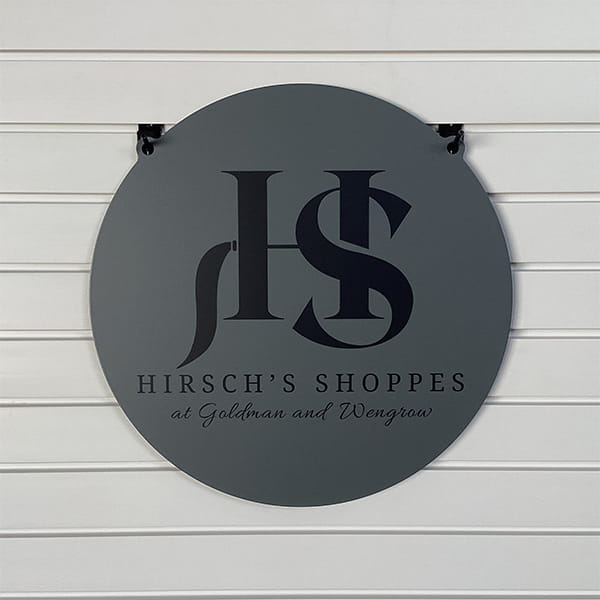 Round metal sign featuring the “Hirsch's Shoppes” logo mounted on a white wall, highlighting bold lettering and a clean, professional design.