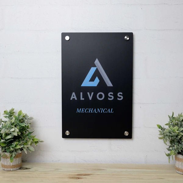 Metal plaque featuring the Alvoss Mechanical logo mounted on a black background, displayed against a white brick wall with plants on either side, highlighting a sleek and professional design