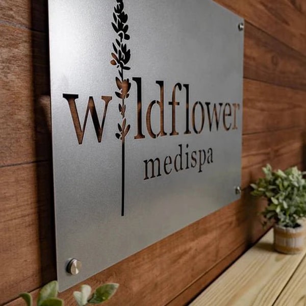 Custom laser-cut metal business sign with "Wildflower Medispa," decorative outdoor wall art mounted on wooden wall, powder coated plaque
