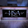 Custom matte black LED backlit metal sign for front door entry, personalized business logo wall art for House Sensational Art Gift Market, modern powder coated outdoor metal signage with warm lighting on wooden counter in dim interior