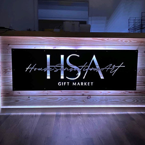 Custom matte black LED backlit metal sign for front door entry, personalized business logo wall art for House Sensational Art Gift Market, modern powder coated outdoor metal signage with warm lighting on wooden counter in dim interior