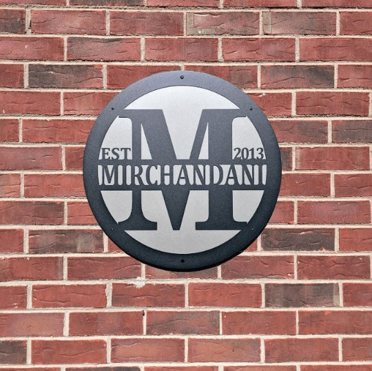 Custom metal gate sign with “Mirchandani” branding, circular personalized family or business décor mounted on brick wall exterior.