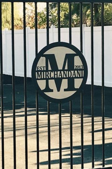 Custom metal gate sign with “Mirchandani” logo and text, personalized family or business décor on gate with white fence and greenery background.