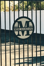 Custom metal gate sign with “Mirchandani” logo and text, personalized family or business décor on gate with white fence and greenery background.