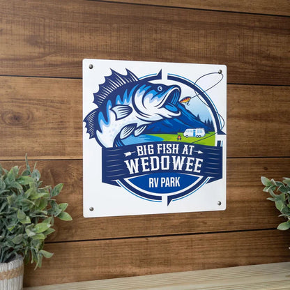 White sign with navy blue text and leaping fish graphic reads "BIG FISH AT WEDOWEE RV PARK," with scenic background and RV.