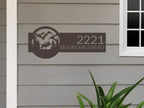 House number sign with mountain and landscape design on gray siding wall, rustic outdoor moose cabin decor