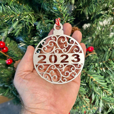 Decorative Christmas ornament displaying “2023” held in front of a Christmas tree, with soft green branches and festive lights in the background creating a warm holiday atmosphere.