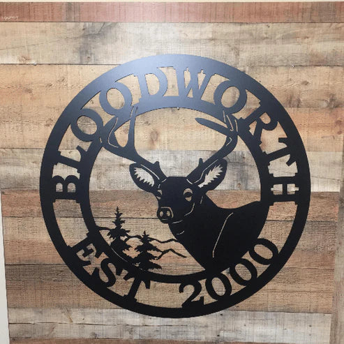 Black circular metal sign with cut-out deer in center, “BLOODWORTH” curved at top, “2000 EST” below, on vertical wood planks.