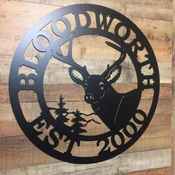 Close-up black circular sign with deer cut-out; “BLOODWORTH” arcs at top, “2000 EST” at bottom, against vertical wood planks.