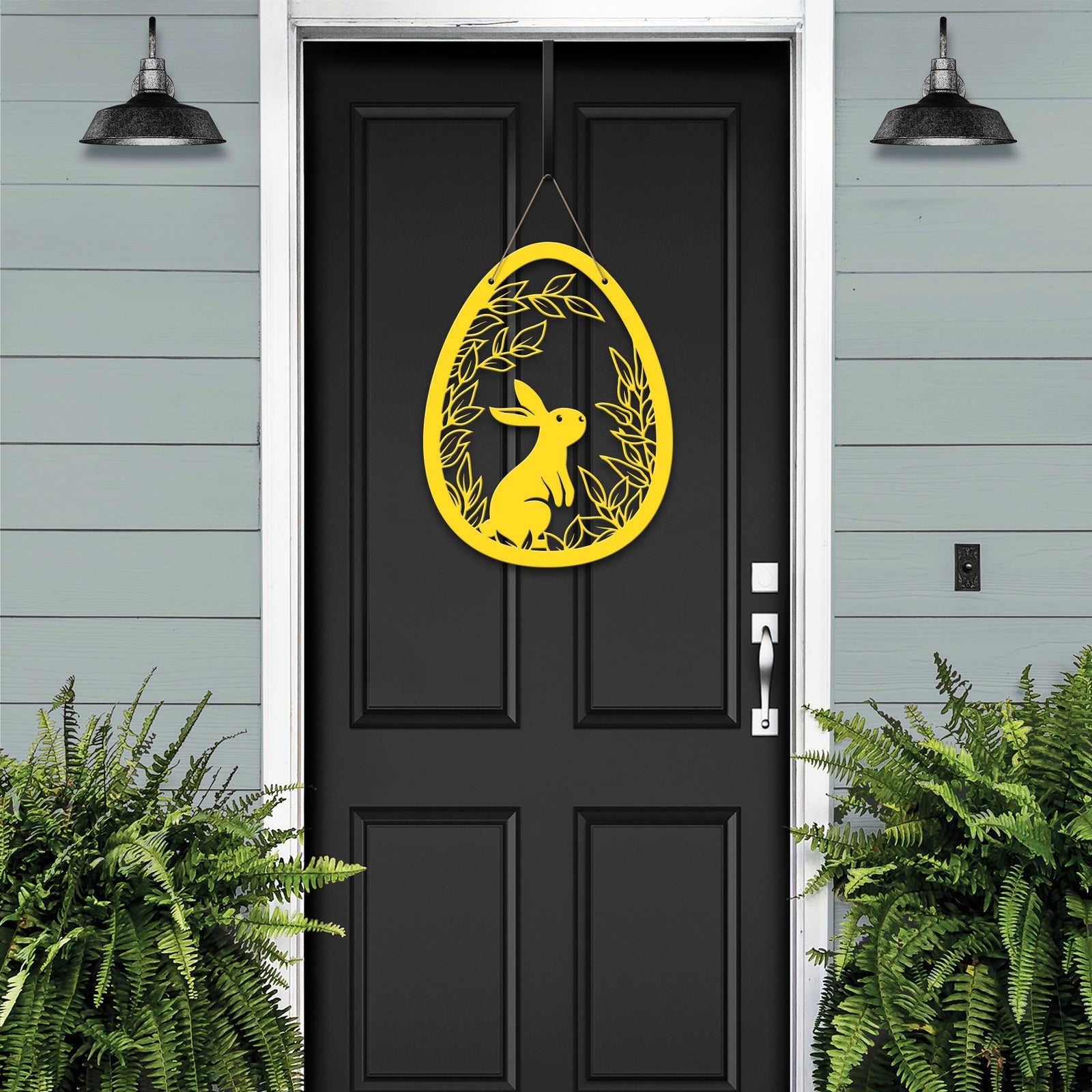 Black door with yellow metal Easter egg door hanger featuring rabbit and leaf cutouts, custom outdoor powder coated spring décor, farmhouse front door sign on light blue wall