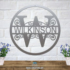 Personalized metal monogram sign featuring the last name “WILKINSON,” displayed on a white brick wall backdrop with two potted plants on either side, highlighting custom craftsmanship and stylish home décor.