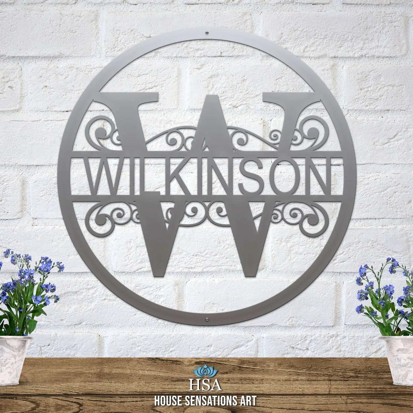 Personalized metal monogram sign featuring the last name “WILKINSON,” displayed on a white brick wall backdrop with two potted plants on either side, highlighting custom craftsmanship and stylish home décor.
