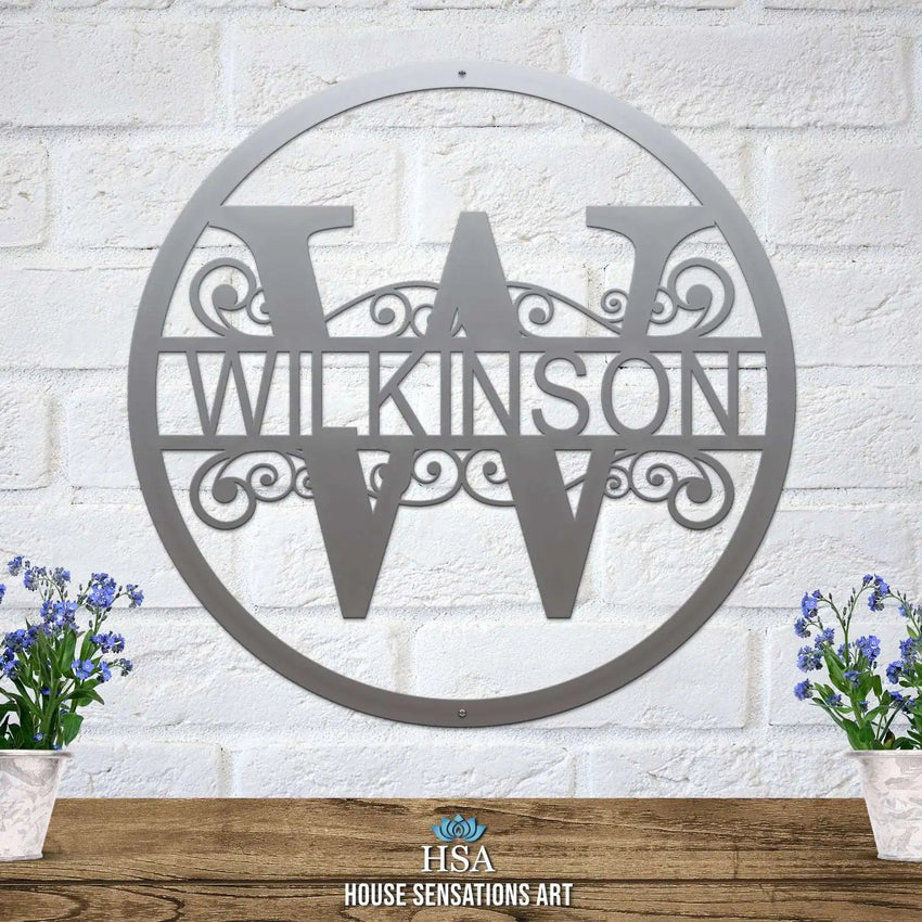 Personalized metal monogram sign featuring the last name “WILKINSON,” displayed on a white brick wall backdrop with two potted plants on either side, highlighting custom craftsmanship and stylish home décor.