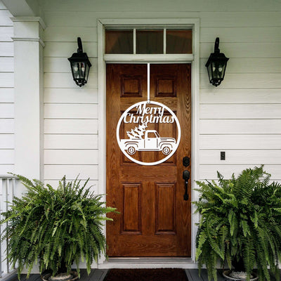 Wooden front door with Merry Christmas truck and tree design, flanked by two potted ferns on white house exterior, festive holiday home décor and seasonal entrance display