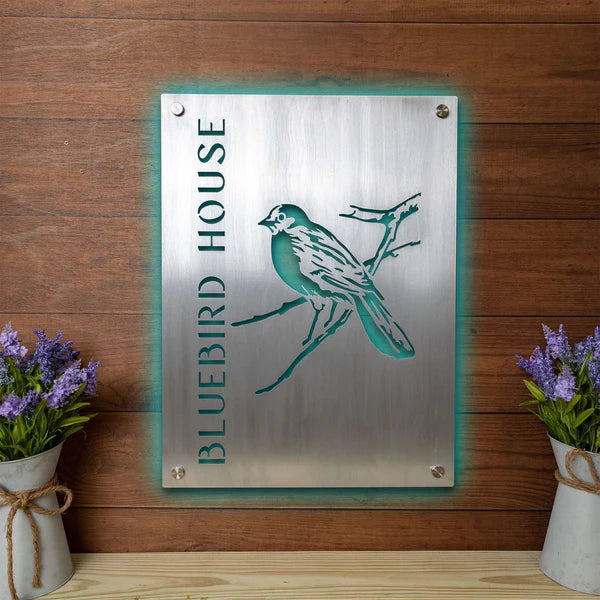 Aluminum sign laser cut and engraved, turquoise backlit, mounted on wood, with vertical “BLUEBIRD HOUSE” and bird on branch design.