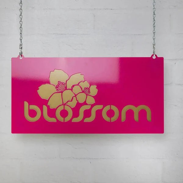 Pink decorative sign with floral design and text “Blossom” hanging on light gray wall, custom indoor or outdoor home décor accent