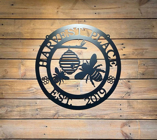 Black laser cut ranch sign with bee silhouettes, beehive, honeycomb accents, and “HARVEST PLACE EST 2019” text; mounted on wood panel, garden decor.