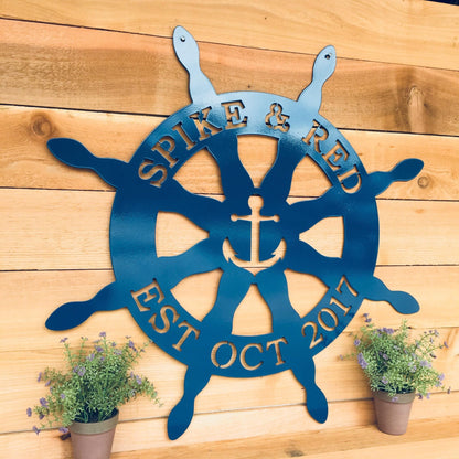 Nautical Captain's Ship Wheel Metal Door Hanger displayed on a wooden wall, surrounded by potted plants, showcasing its customizable design and sturdy craftsmanship ideal for beach house decor.