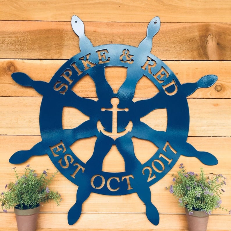 Nautical Captain's Ship Wheel Metal Door Hanger displayed on a wooden wall, accompanied by potted plants with purple flowers, showcasing its suitability for beach house personalization.