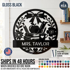 Introducing the Personalized Teacher Name Classroom Sign - a round custom metal sign shown in Satin Black displayed on a homeschool classroom wall. Features a beautiful collage of schoolbooks, teacher's apple, ruler, pencil in coffee mug, and ornate bow & flower, with personalized name plaque across the lower half. Featured photo reads Mrs. Taylor. Sign comes with color-matching screws and is available in more than 50 color finishes