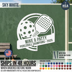 A freeform custom metal sign with pickleball paddle & pickleball plus personalized names shown in Sky White on green pickleball paddle court wall. Sign ships in 48 hours or less when ordered before noon, making it a fast couples gifts. Included free color-matching screws for ready-to-mount gifting. - ideal for retirement gift for any pickleball loving couple.
