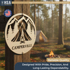 Round metal custom camper sign with mountain and pine tree cutouts, reads CAMPER VILLE, mounted on a post at a sunlit campsite.