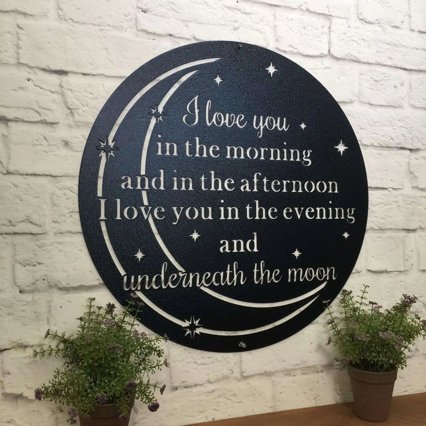 Custom Metal Quote Sign displayed on a wall, featuring personalized text. Accompanied by potted plants, showcasing its suitability for home, office, or outdoor décor. Made from durable 14-gauge steel.