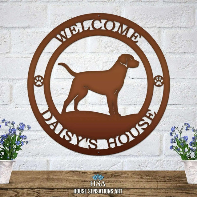 A candy copper metal wall sign showing a Labrador dog silhouette standing, with “WELCOME,” “DAISY'S HOUSE,” and paw prints on each side.