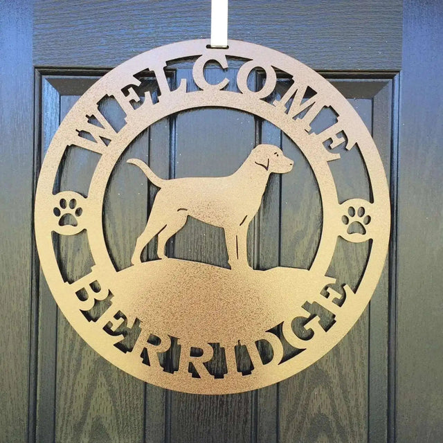 A gold metal wall sign showing a Labrador dog silhouette standing, with “WELCOME,” “BERRIDGE,” and paw prints on each side.