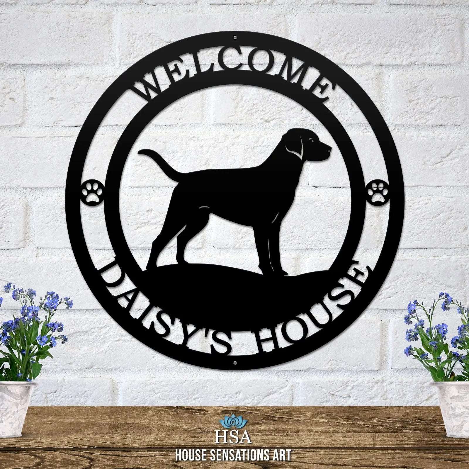 A black metal wall sign showing a Labrador dog silhouette standing, with “WELCOME,” “DAISY'S HOUSE,” and paw prints on each side.