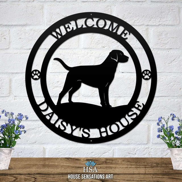 A black metal wall sign showing a Labrador dog silhouette standing, with “WELCOME,” “DAISY'S HOUSE,” and paw prints on each side.