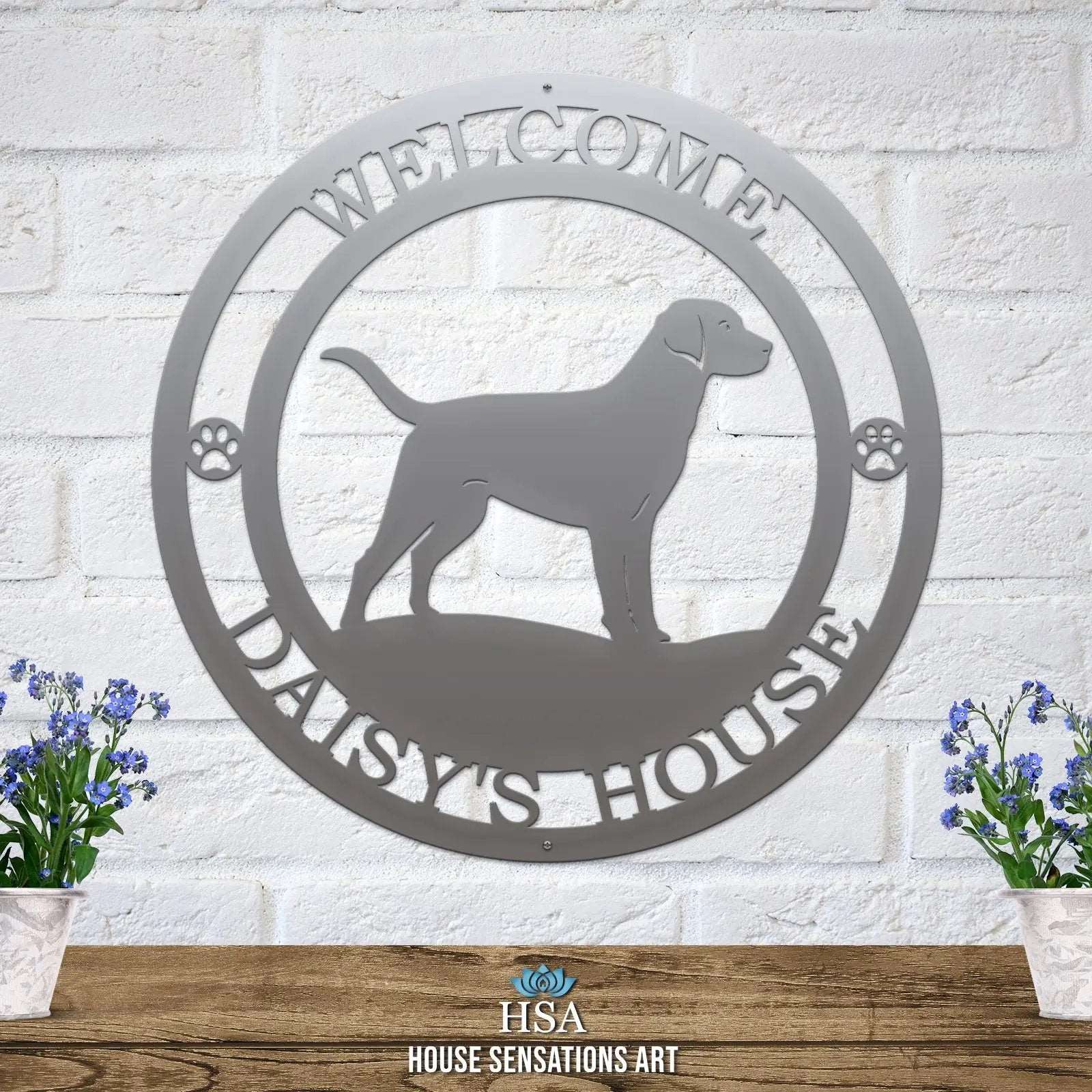 A polished chrome metal wall sign showing a Labrador dog silhouette standing, with “WELCOME,” “DAISY'S HOUSE,” and paw prints on each side.