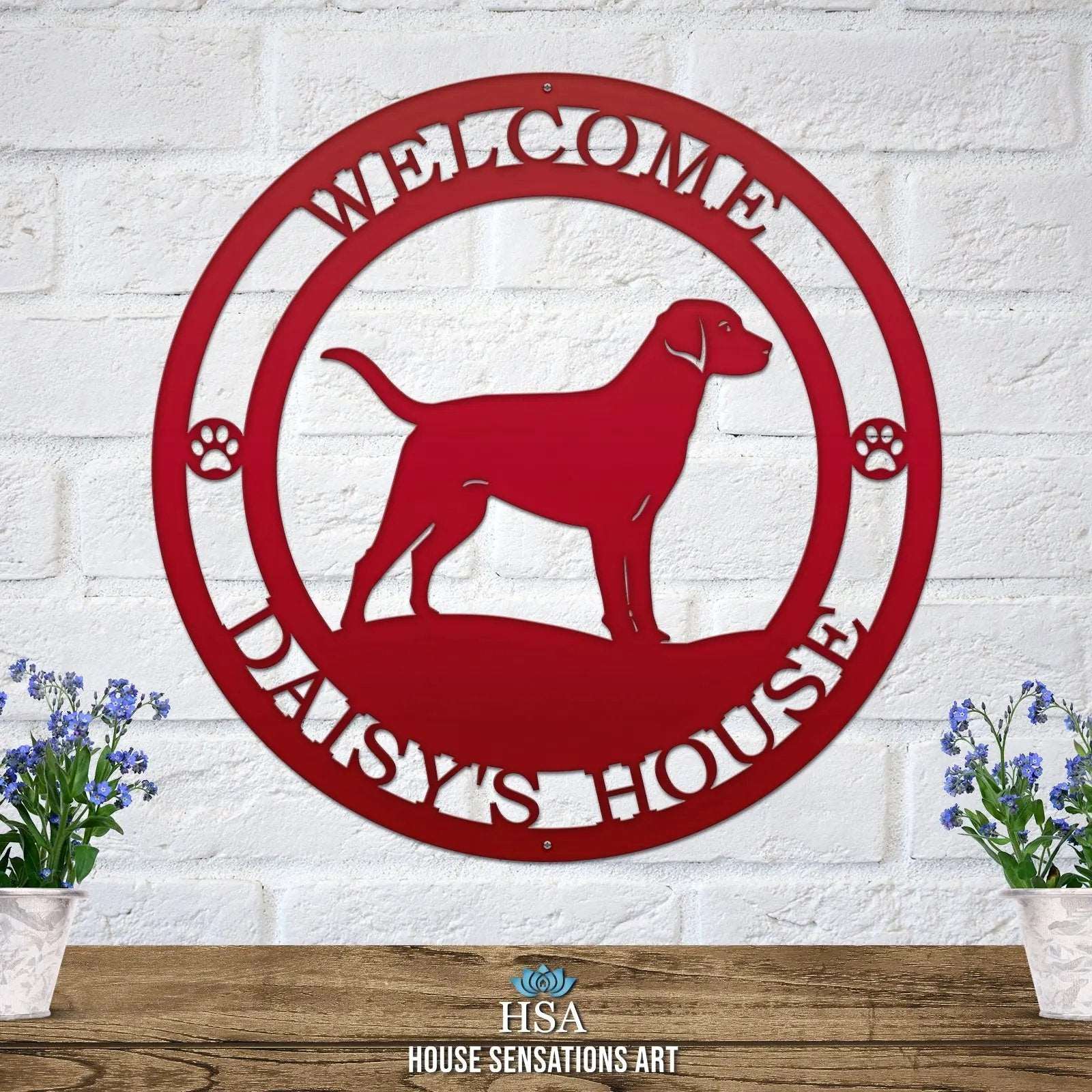 A deep red metal wall sign showing a Labrador dog silhouette standing, with “WELCOME,” “DAISY'S HOUSE,” and paw prints on each side.