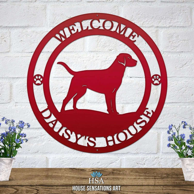 A deep red metal wall sign showing a Labrador dog silhouette standing, with “WELCOME,” “DAISY'S HOUSE,” and paw prints on each side.