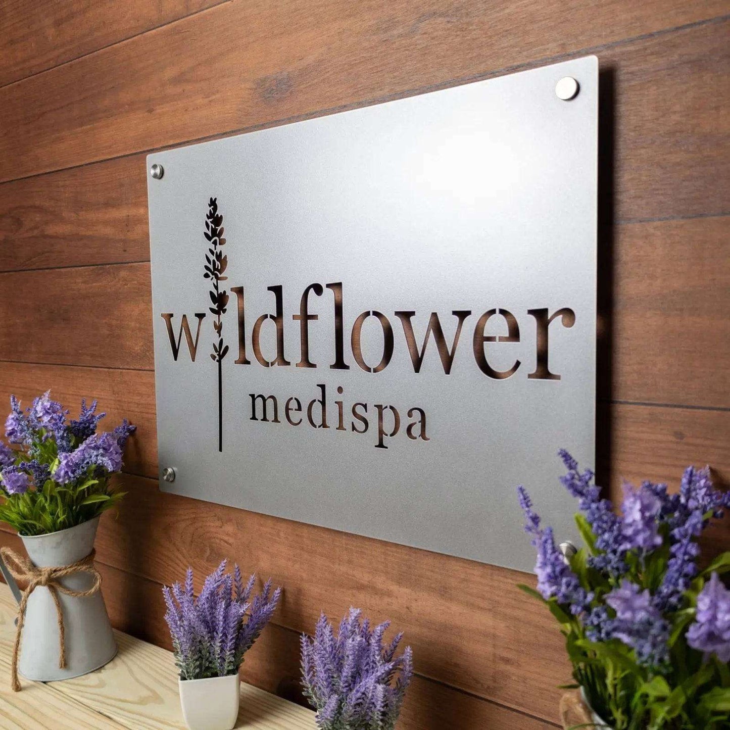 Large custom laser-cut metal business logo sign for “Wildflower Medispa” displayed on a wooden wall with lavender plants, showcasing personalized office or storefront décor from House Sensations Art.