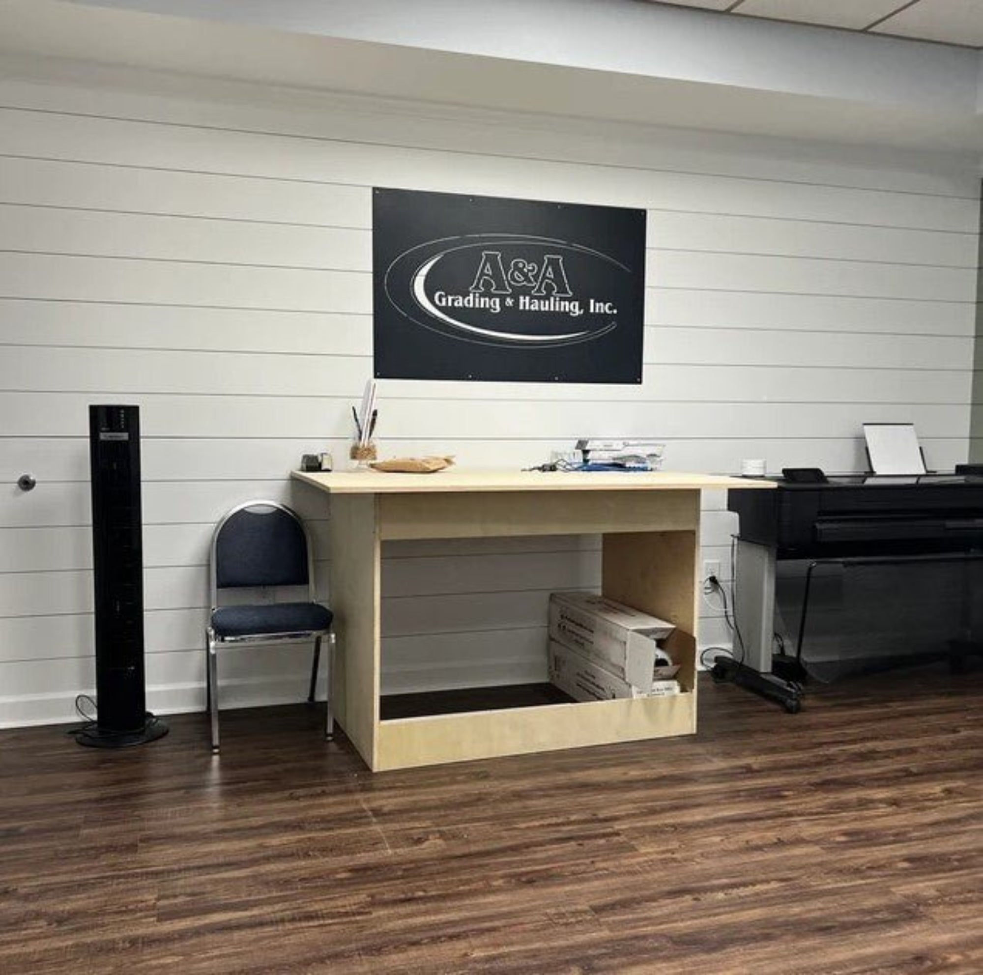 Small office setup with desk, chair, and computer equipment featuring a large custom laser-cut metal sign of "A&A Grading & Hauling Inc." mounted on a white wall – House Sensations Art.