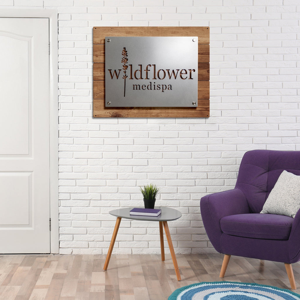 Large custom metal business logo sign for “Wildflower Medispa” mounted on a white brick wall, displayed in a living room with a purple armchair, showcasing personalized laser-cut office or storefront décor.