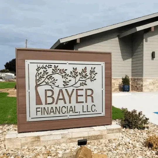 Custom outdoor metal business sign for “Bayer Financial, LLC” — laser-cut aluminum with tree branch detail, mounted in a wooden frame on a stone base.