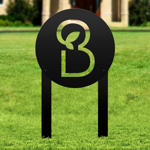 Laser-cut black metal sign with “B” and leaf in circular frame, mounted on two legs, placed in grassy outdoor area.