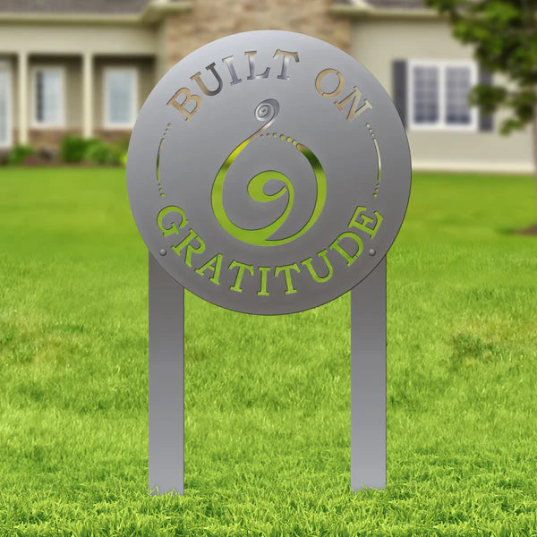 Polished silver laser-cut sign with spiral and “BUILT ON GRATITUDE,” mounted on posts in grass with house in background.