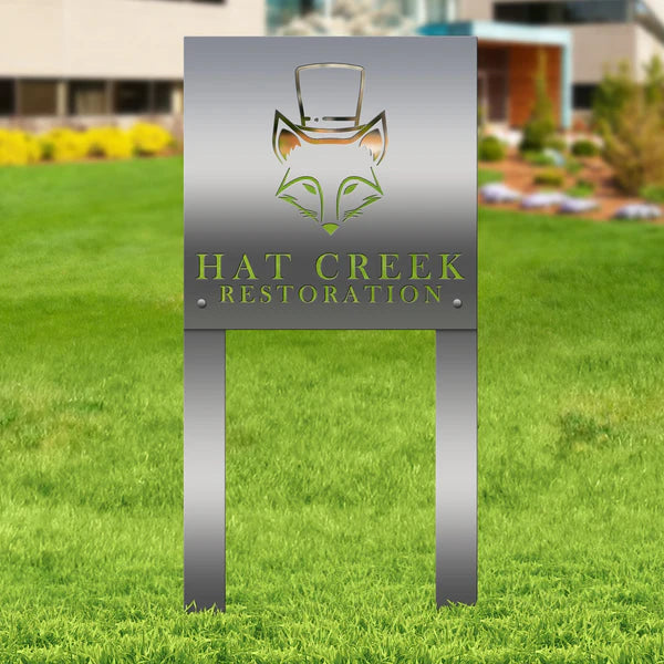 Polished silver metal sign with fox in top hat and “HAT CREEK RESTORATION,” mounted on posts in grassy landscaped area.