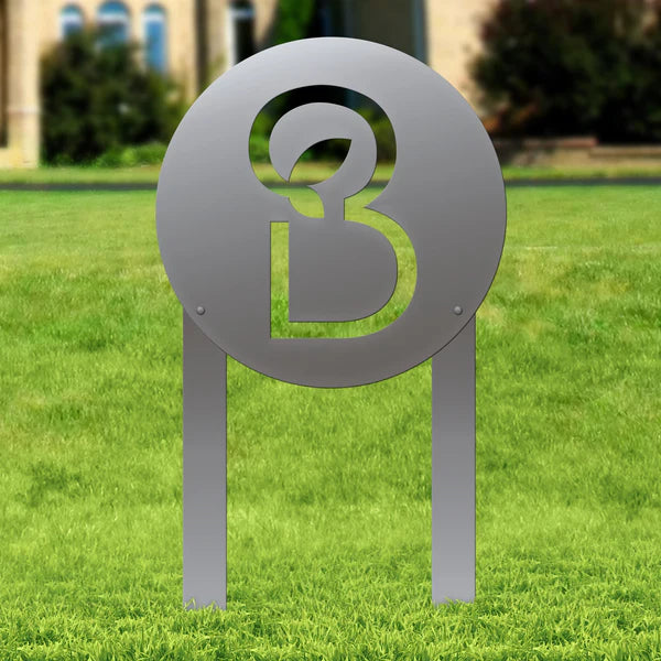 Polished silver sign with circular frame and abstract “B” design, standing on two posts in grass with soft landscaping behind.