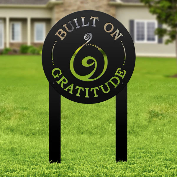 Black laser-cut sign with “BUILT ON GRATITUDE” and spiral cutout, mounted on posts in grass lawn with house visible behind.