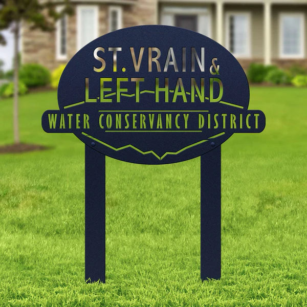 Textured navy oval sign with “ST. VRAIN & LEFT HAND WATER CONSERVANCY DISTRICT,” mounted in grass with building behind.