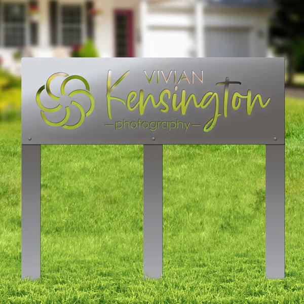 Polished silver metal sign with floral design and “VIVIAN Kensington photography,” mounted on posts in grass with building behind.