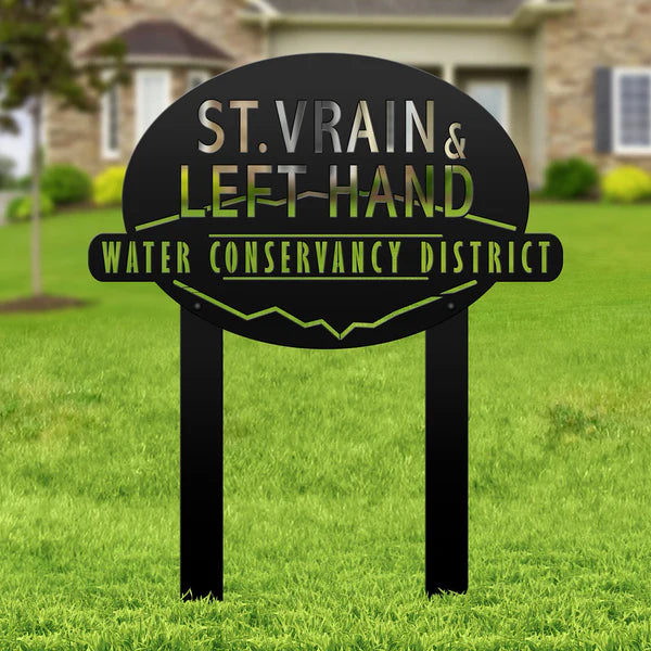 Black oval sign with “ST. VRAIN & LEFT HAND WATER CONSERVANCY DISTRICT,” mounted in grass with building behind.